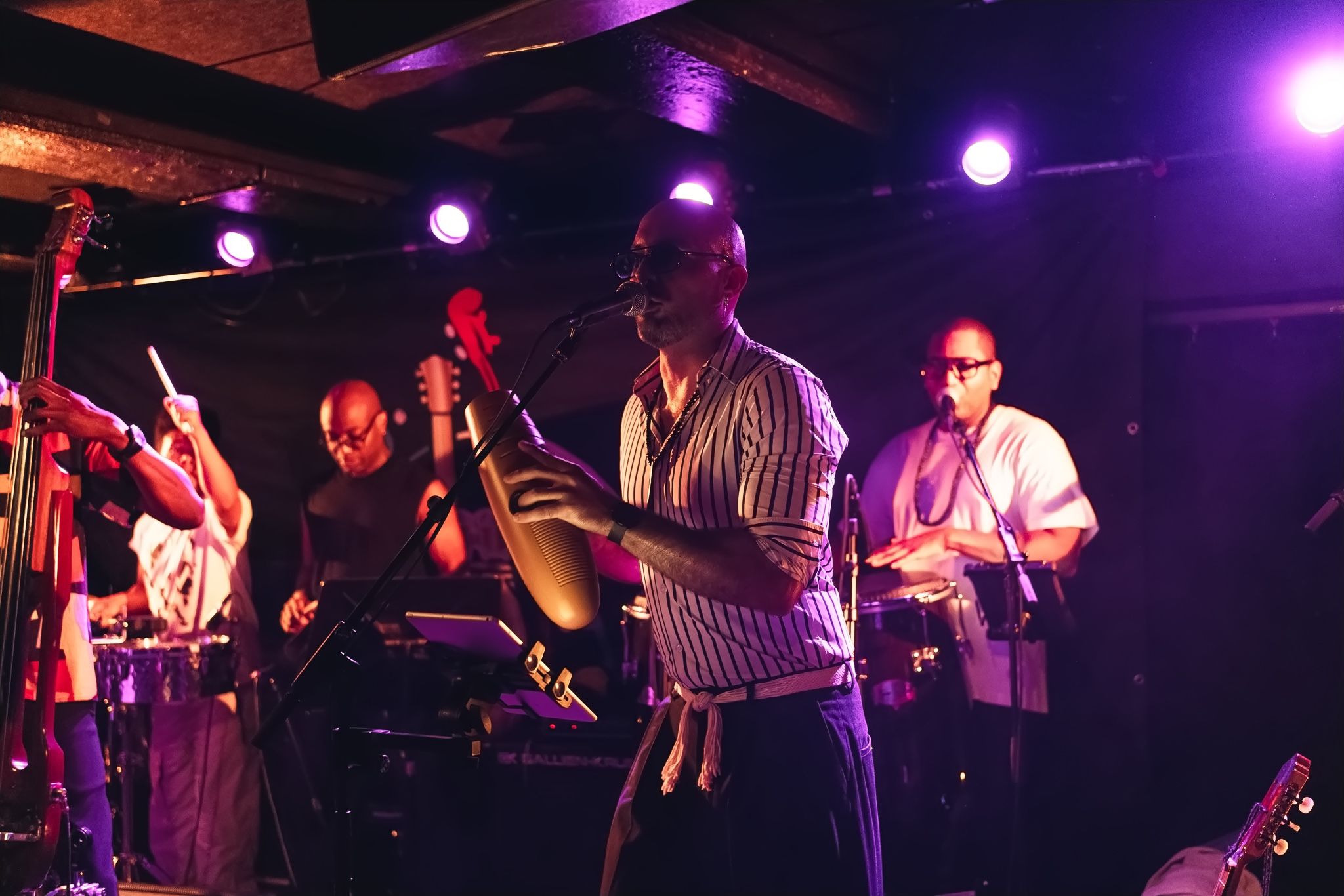 Singer with maracas in front of the bass, timbales and congas under violet light.