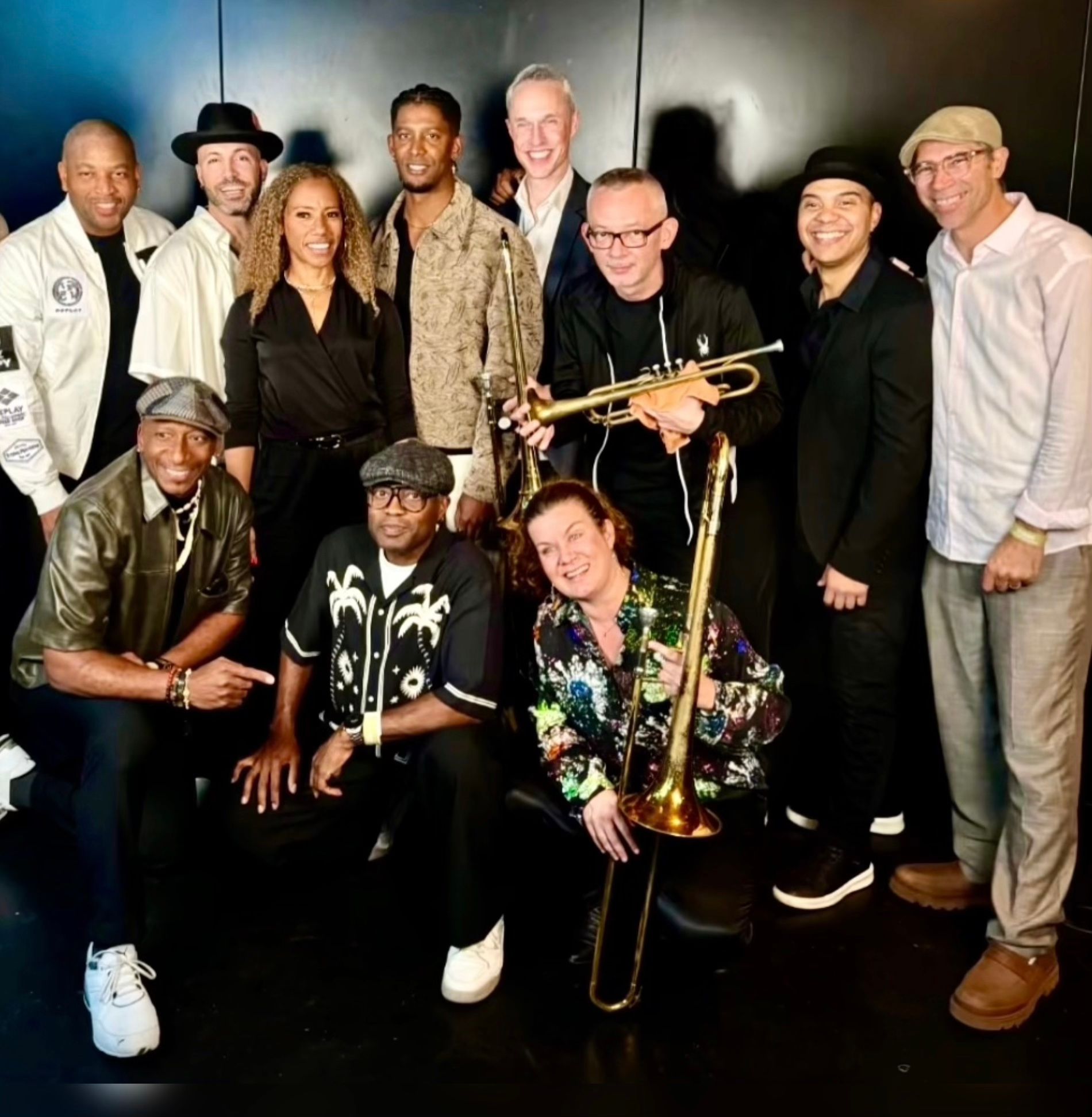 The musicians of Eminente All Stars posing together against a dark wall, three kneeling in front, one holding a trumpet and one a trombone.